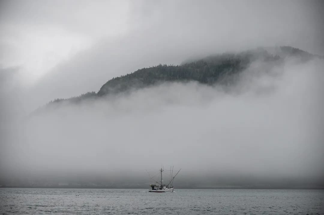 Small fishing vessel crossing fog-wrapped water, forested mountain ridge emerging through low clouds above.