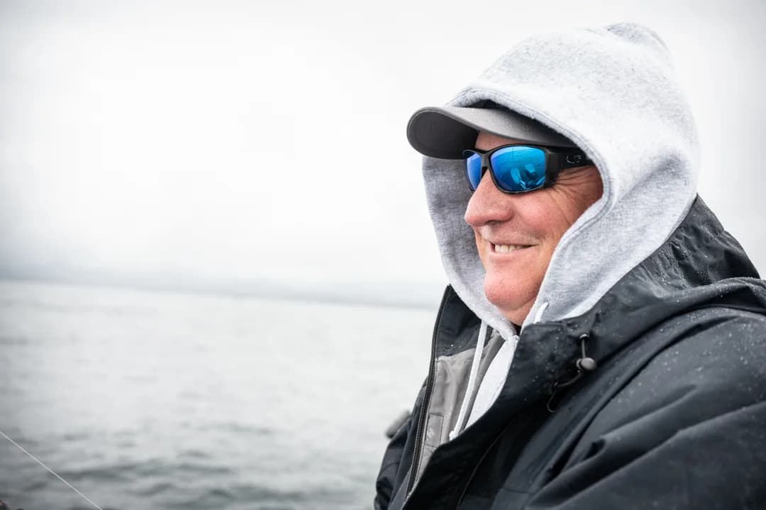 Angler in rain gear and polarized sunglasses smiling on the water, overcast skies behind him.