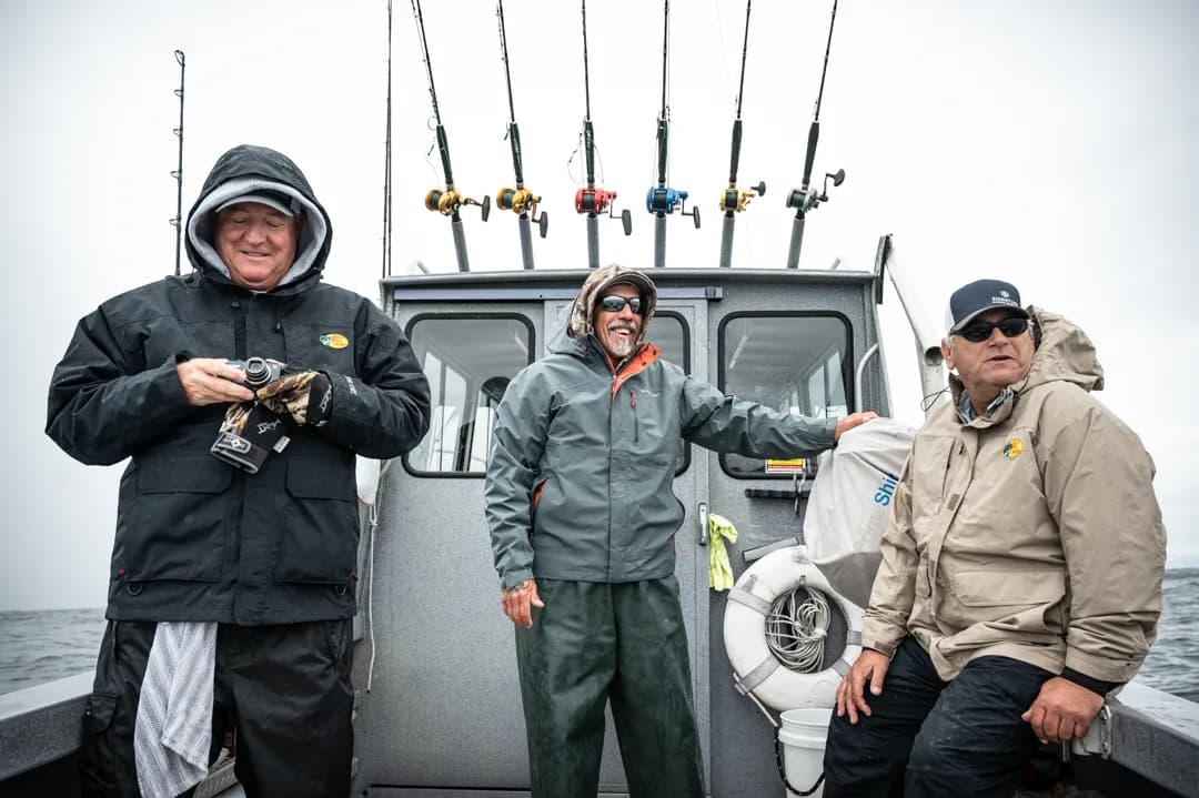Three anglers in rain gear on the stern deck, rod rack and aluminum wheelhouse behind them, overcast open water.