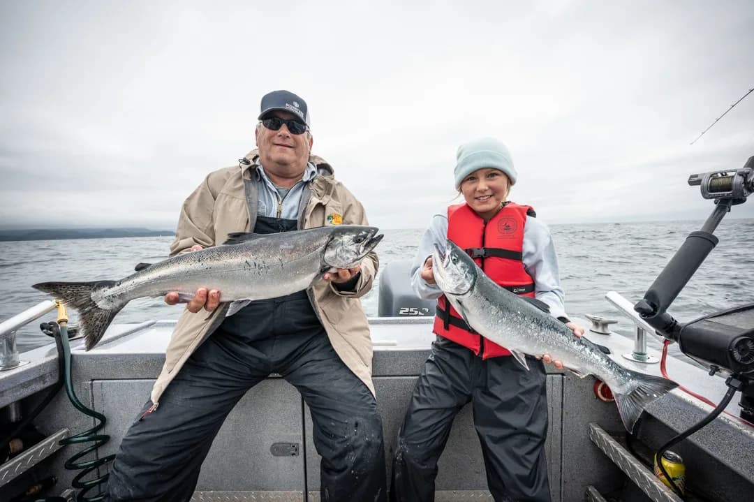 Man and young girl each hold a silver salmon on the deck of an aluminum fishing boat, overcast sky and open water behind them.