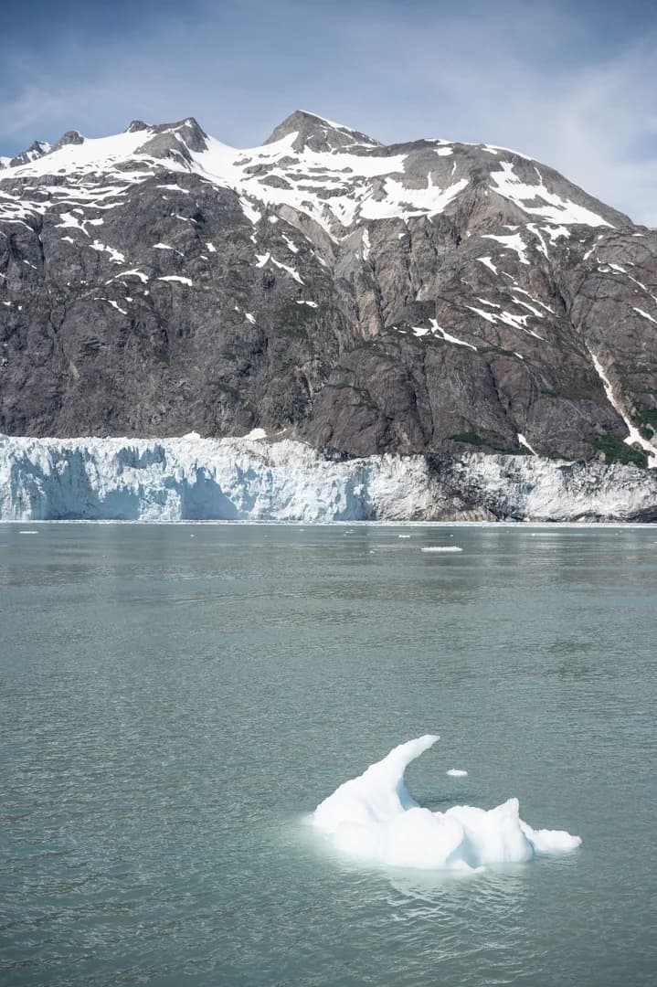 Small iceberg floating in front of a tidewater glacier face, snow-capped mountains rising behind, Glacier Bay, Alaska.