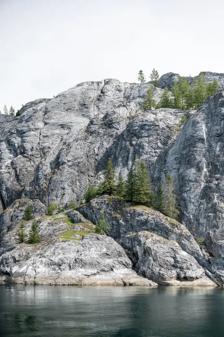 Glacier-polished granite cliffs rise from calm dark water, sparse conifers clinging to ledges above the shoreline.