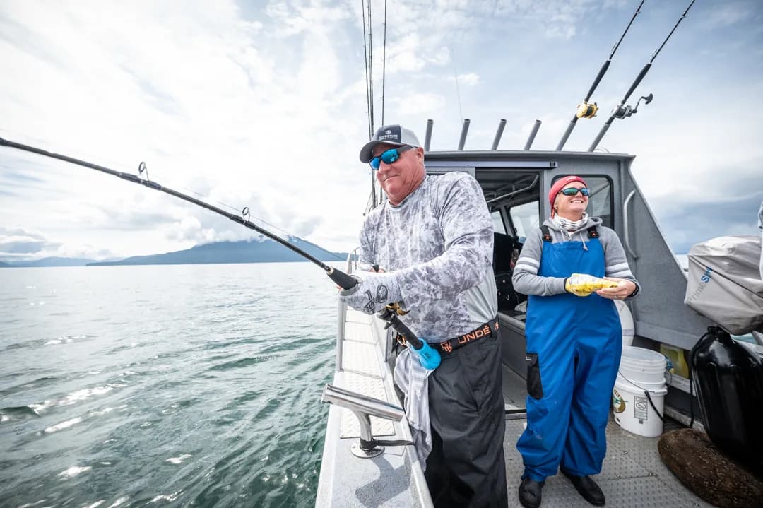 Angler working a bent rod on the stern of a charter boat, guide in blue bibs looking on, mountains across the water.