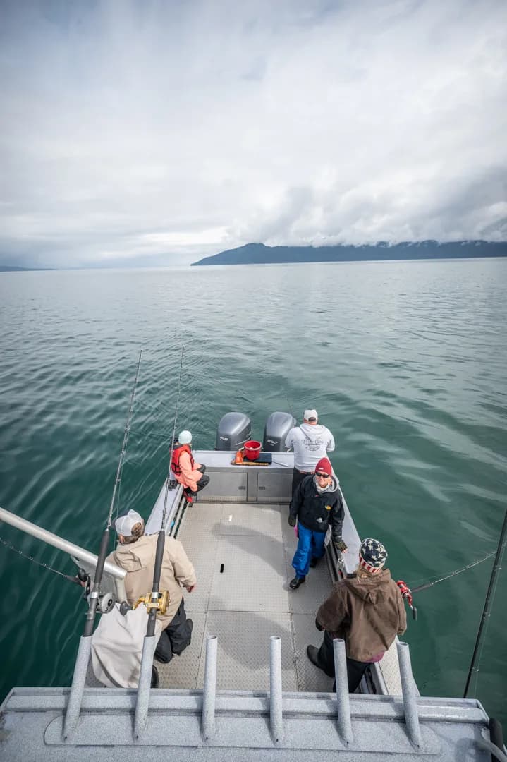 Anglers on the stern deck of an aluminum fishing boat, rods deployed, cloud-draped coastal mountains in the distance.
