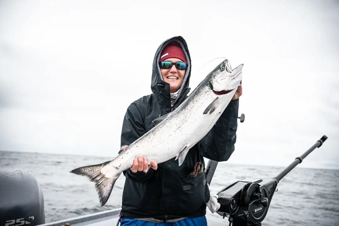 Angler in rain gear holds a silver salmon on an overcast day on the water, rod holder and electronics visible behind her.