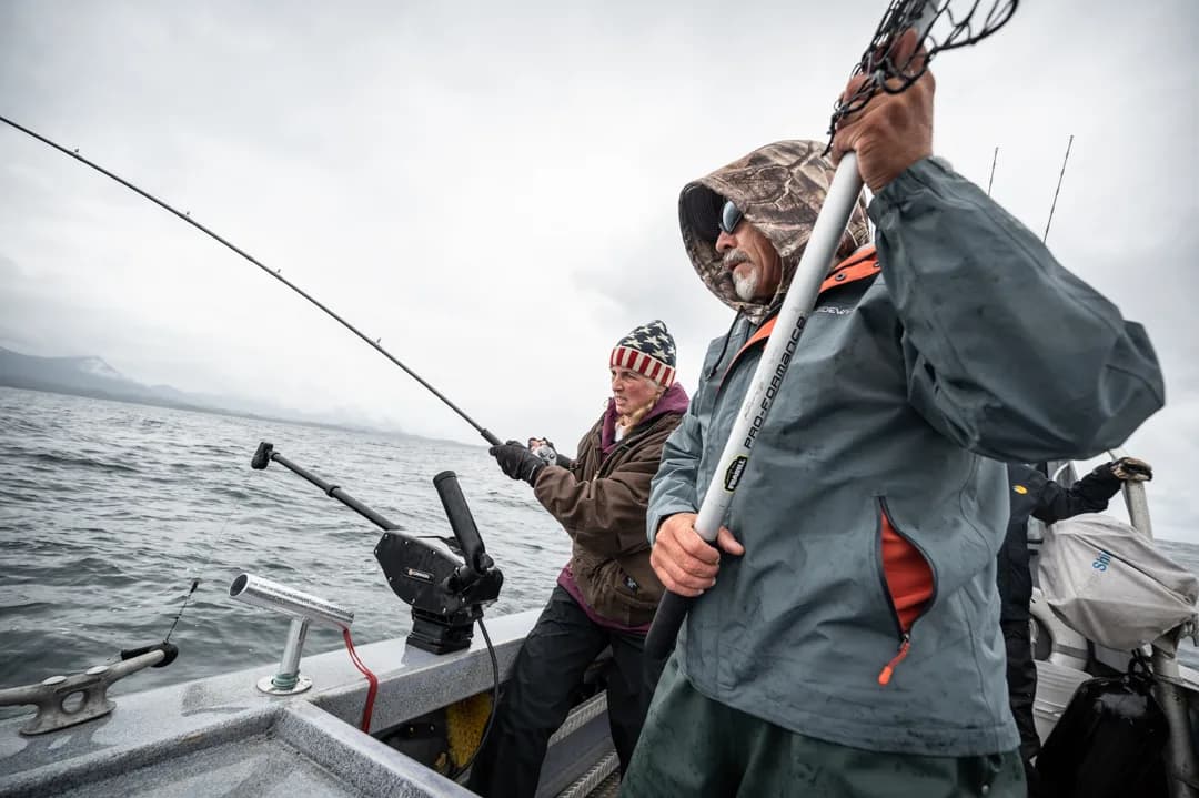 Two anglers working rods on an aluminum boat deck in overcast conditions, mountains visible in the mist behind them.