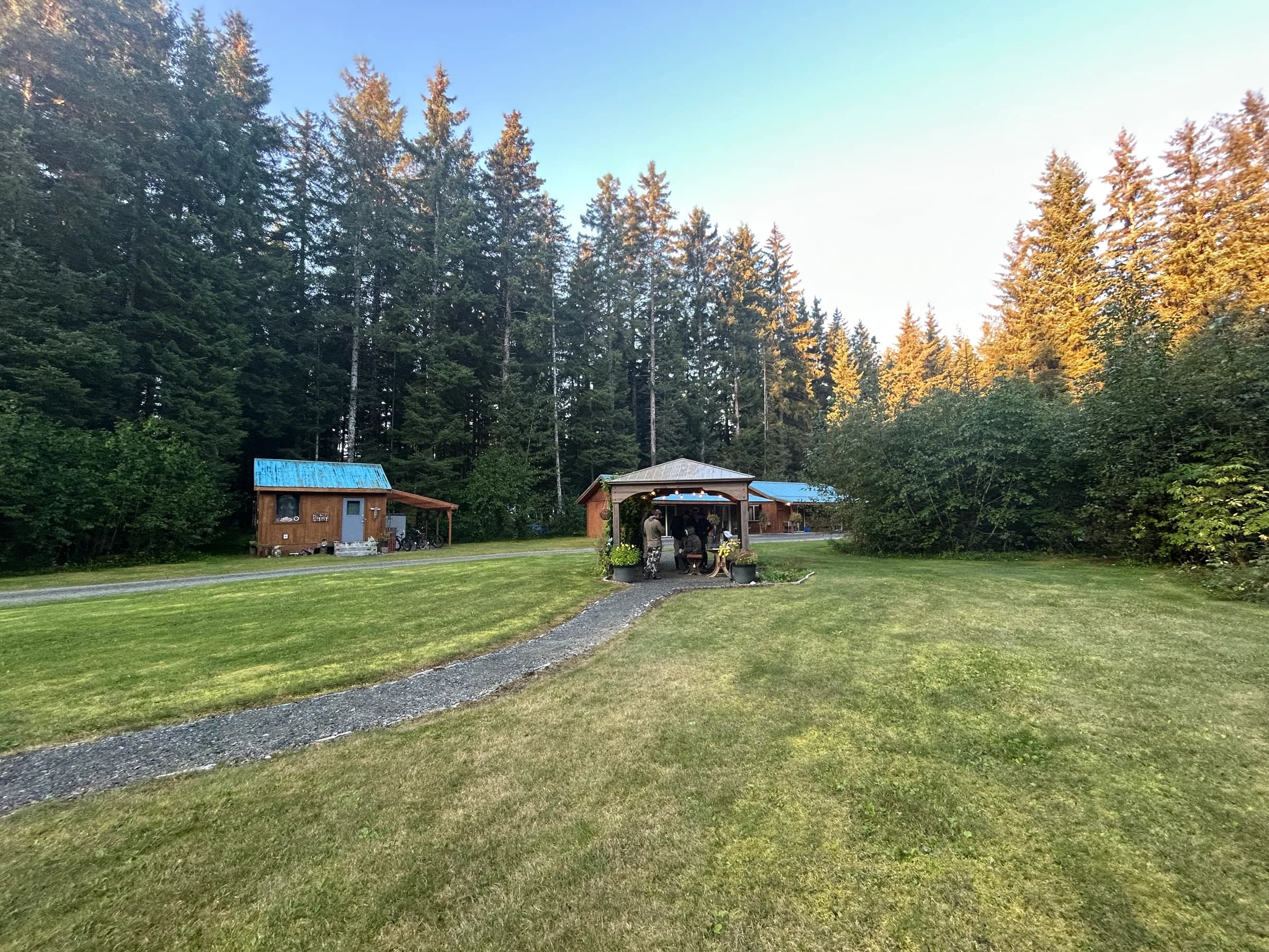 Lodge entry structure and outbuilding on a gravel path, surrounded by spruce forest in warm evening light, Gustavus Alaska.