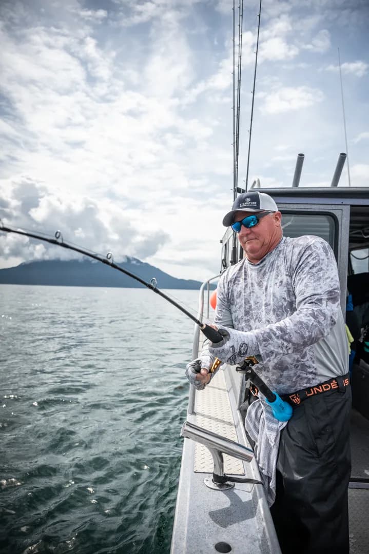 Angler working a bent rod off the stern rail, mountains and open water visible behind him.