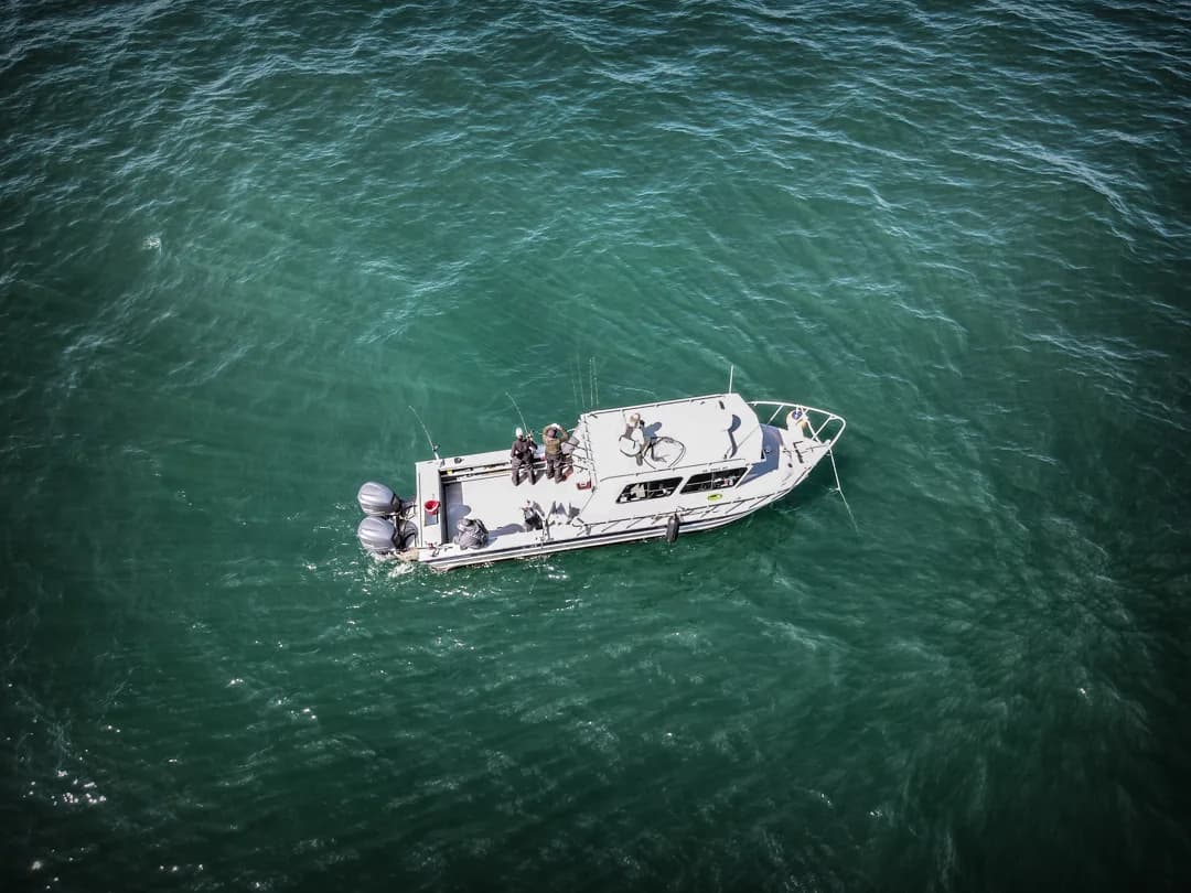 Aerial view of a white sport-fishing boat with anglers on deck, drifting over teal-green water.