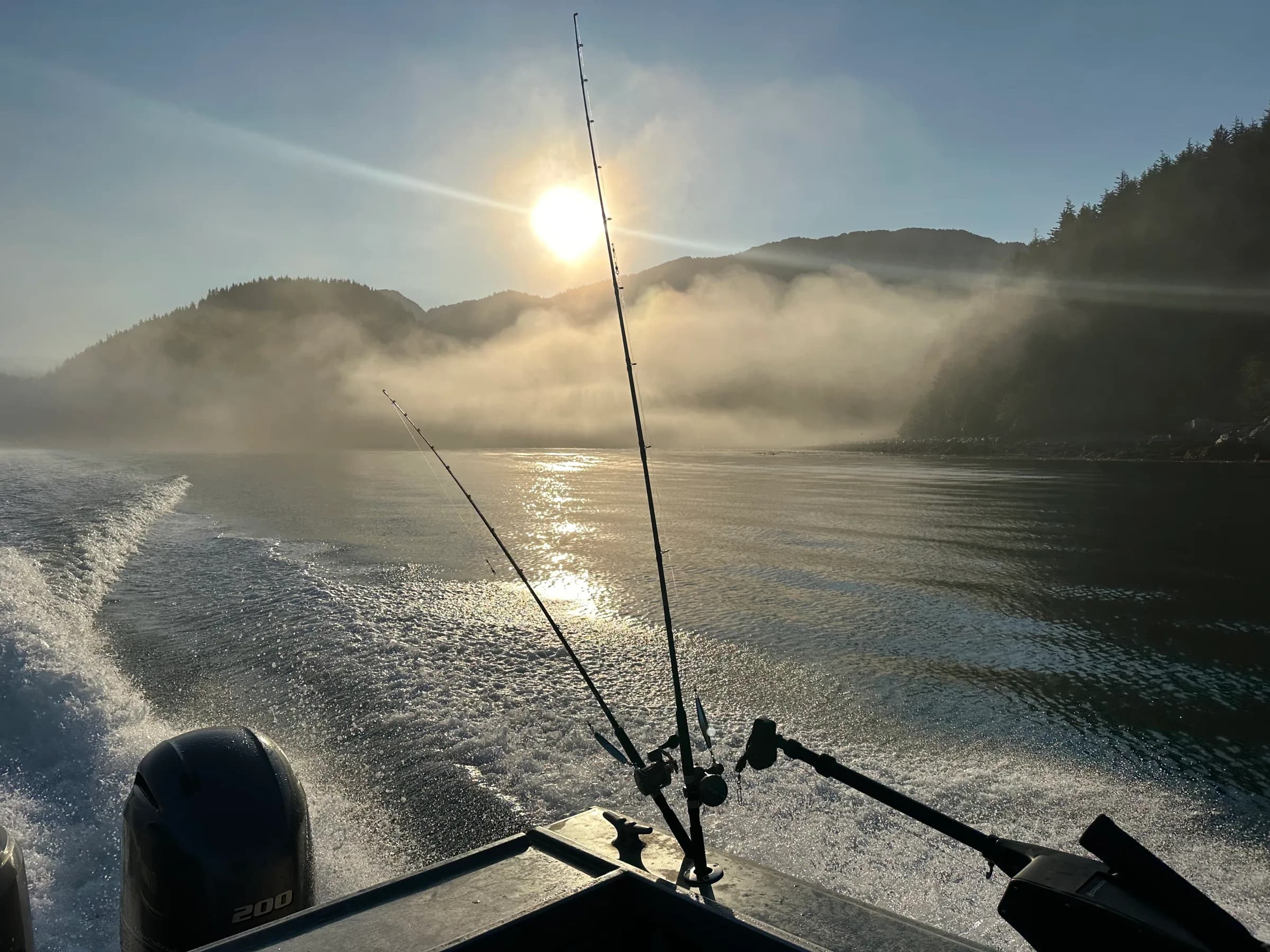 Fishing rods silhouetted against morning sun and low mist over mountain-flanked inlet, wake trailing behind outbound boat.