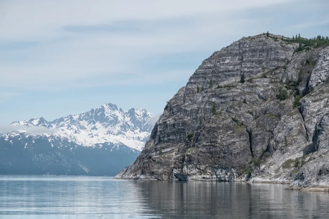 Steep granite cliff rises from calm water in Glacier Bay, snow-capped peaks visible in the distance.