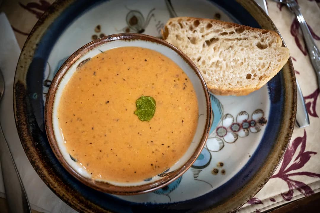Bowl of creamy tomato bisque with herb garnish and crusty bread slice on a floral ceramic plate.