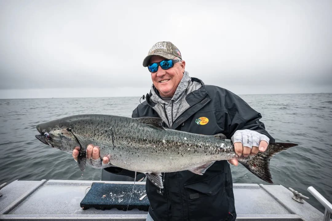 Angler holding a large king salmon on the boat deck, overcast sky and open water behind him.
