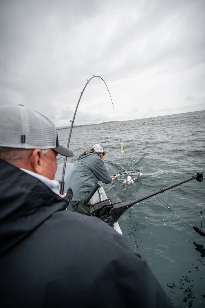 Angler nets a fish at the gunwale while a second rod bends under load, overcast Icy Strait in the background.