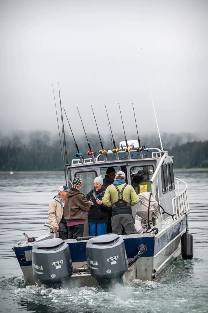Charter fishing boat with twin Yamaha 250 outboards and full rod rack underway on calm gray water, forested shoreline behind.