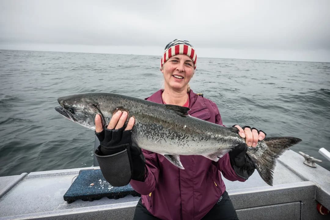 Woman on boat deck holding a silver salmon with open ocean behind her, overcast sky.