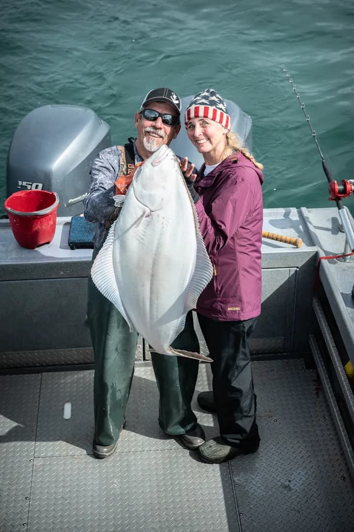 Two anglers holding a large halibut on the deck of an aluminum fishing boat, outboard motors and green water behind them.