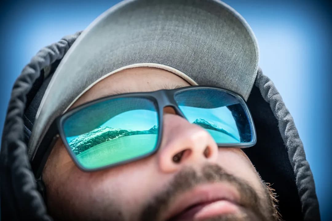 Angler's mirrored sunglasses reflecting snow-capped mountains and glacial water, close-up portrait against blue sky.