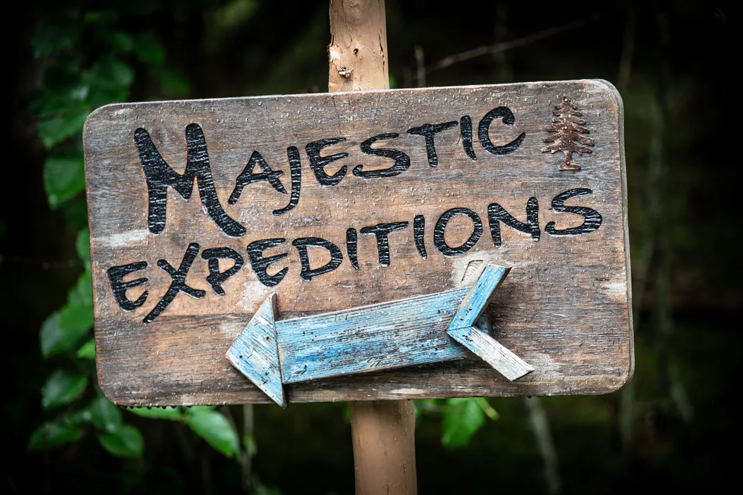 Weathered wooden sign reading Majestic Expeditions with a blue painted arrow, mounted on a post against forest greenery.