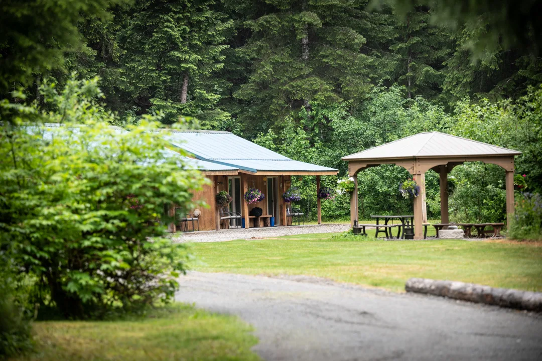 Lodge cabin with blue metal roof and hanging flower baskets beside an open gazebo with picnic tables, set against spruce forest.