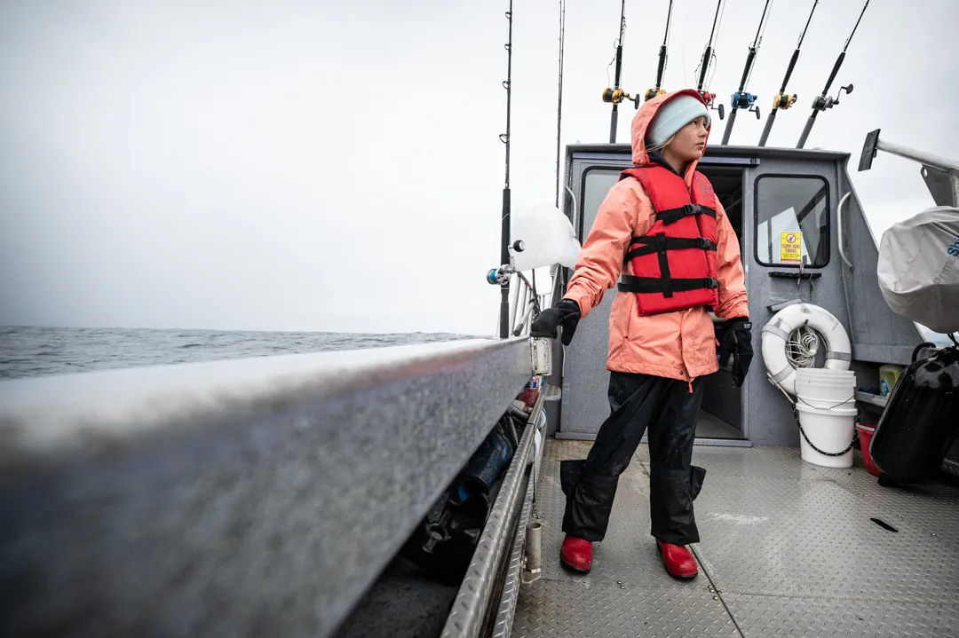 Angler in rain gear and life vest stands on the stern deck of a charter boat, rods racked behind her, gray water stretching to the horizon.
