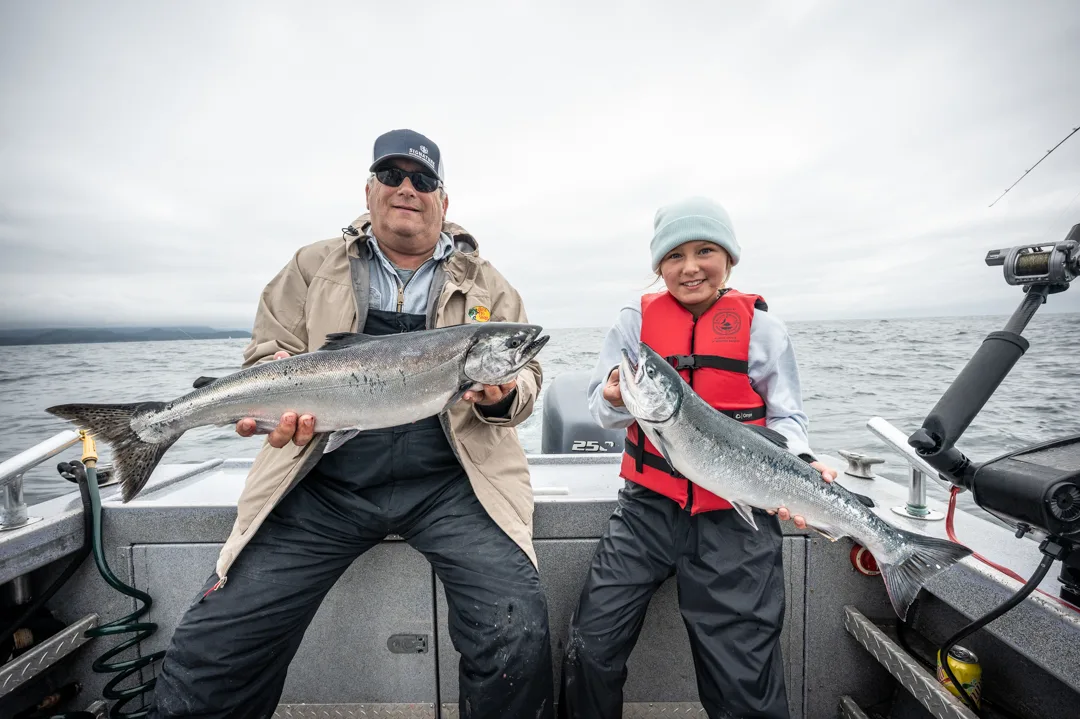 Man and young girl each hold a silver salmon on the deck of an aluminum fishing boat, overcast sky and open water behind them.