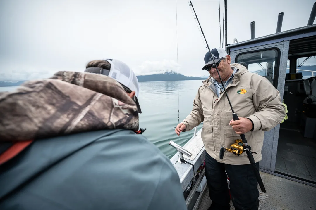 Guide handling terminal tackle on deck while angler watches, snow-capped mountains and calm water behind