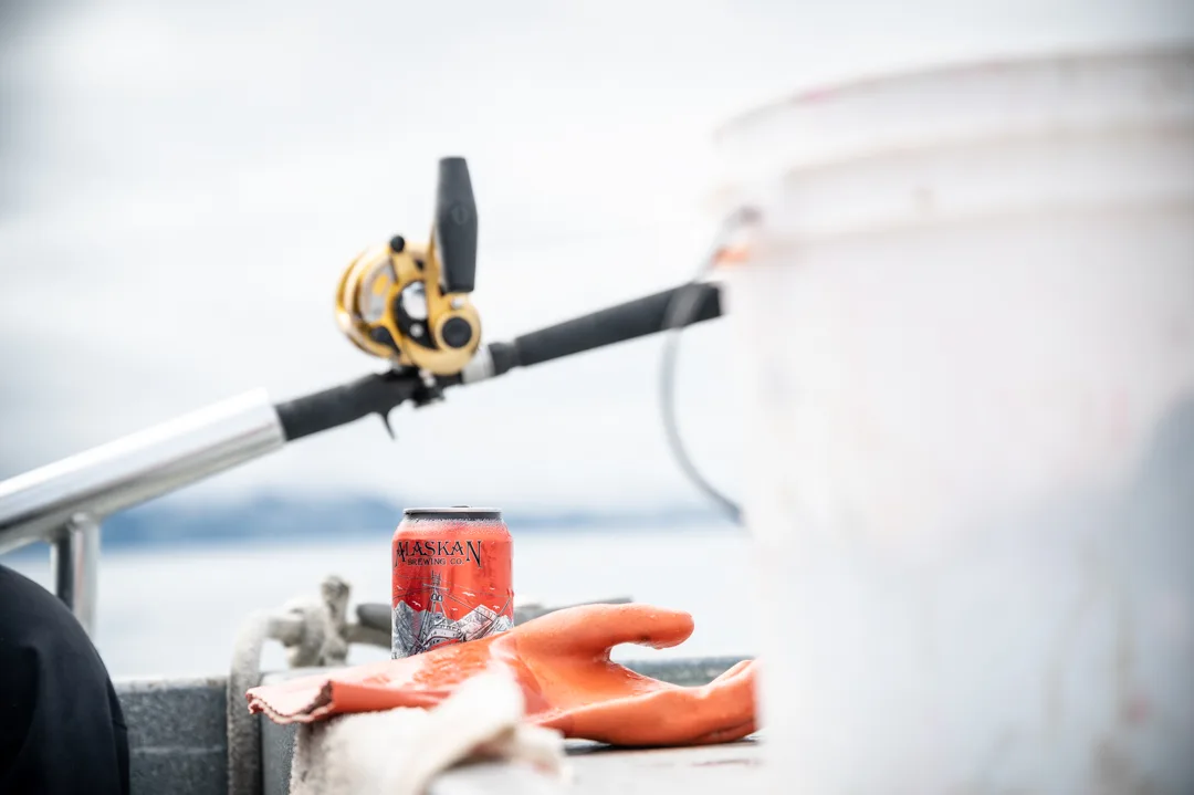 Fishing rod with gold reel resting on boat rail beside an Alaskan Brewing Co. can and orange rubber gloves, water in background.