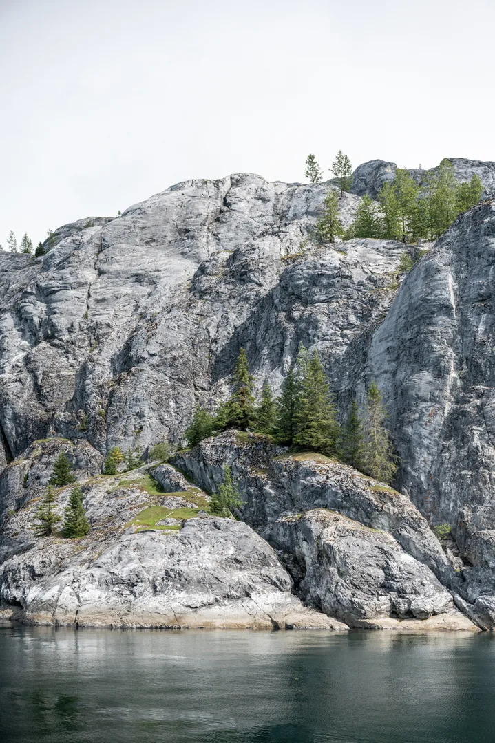 Glacier-polished granite cliffs rise from calm dark water, sparse conifers clinging to ledges above the shoreline.