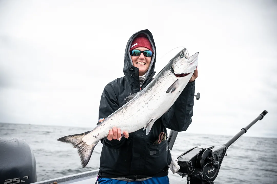 Angler in rain gear holds a silver salmon on an overcast day on the water, rod holder and electronics visible behind her.