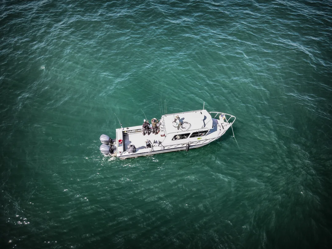 Aerial view of a white sport-fishing boat with anglers on deck, drifting over teal-green water.