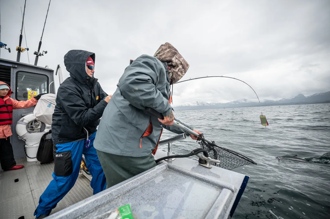 Angler working a landing net over the gunwale as guide assists, rod bent deep, overcast Icy Strait mountains in background.