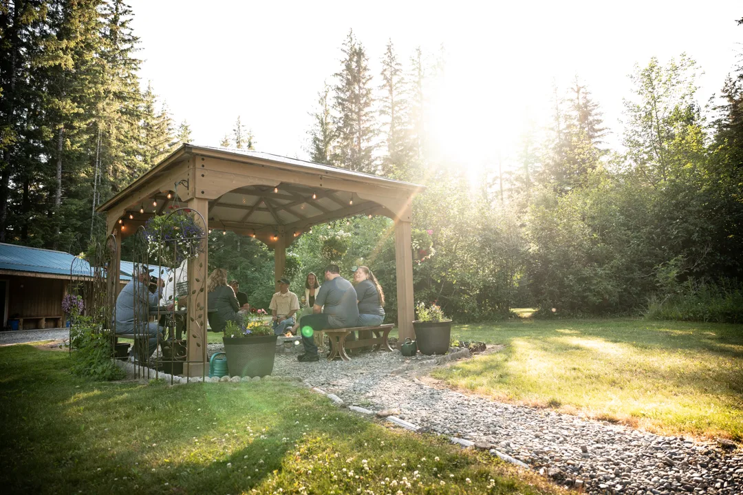 Group of guests gathered under a wood gazebo with string lights at golden hour, lodge building visible at left edge.