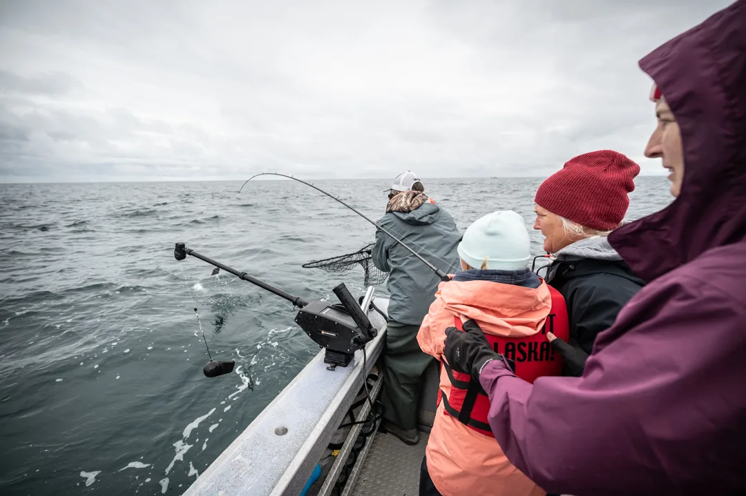 Three anglers lean over the gunwale as a bent rod and landing net work a fish to the surface on overcast open water.