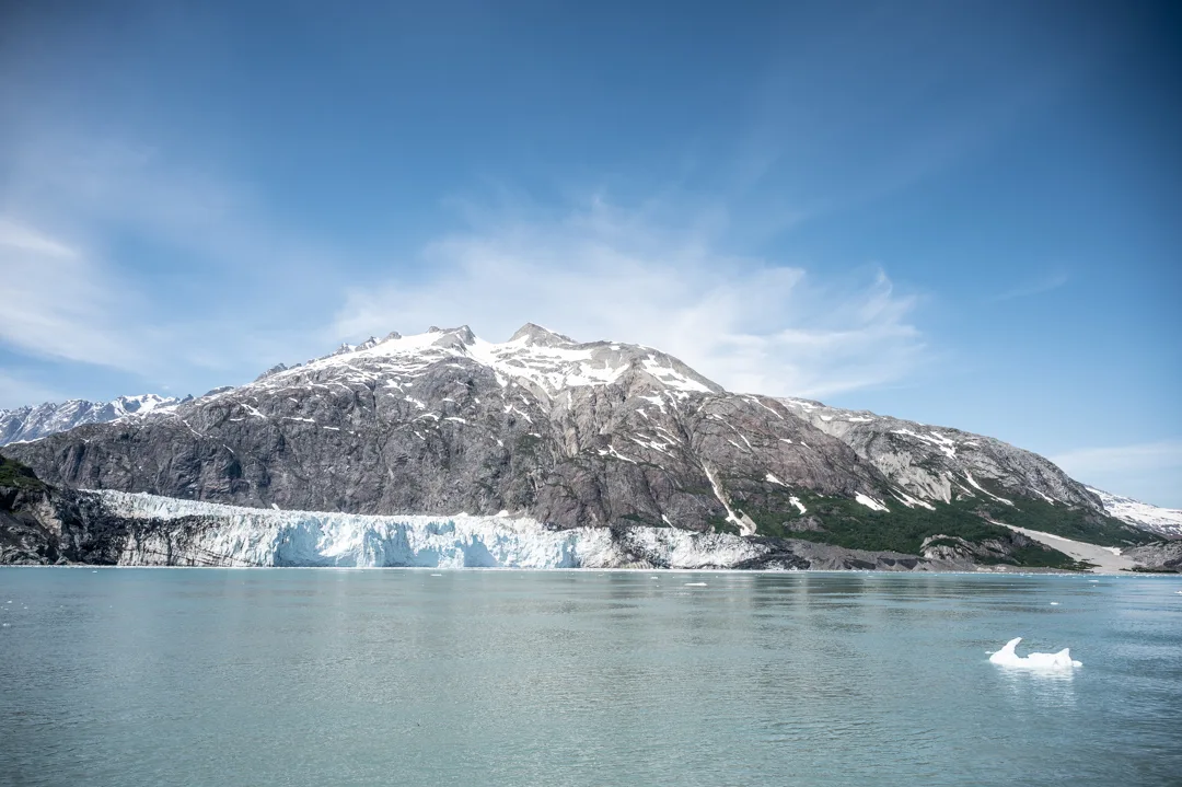 Glacier calving face meets open water in Glacier Bay, small iceberg floating in the foreground, snow-capped peak above.