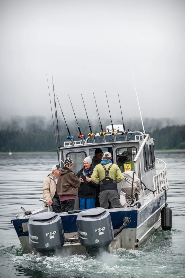Charter fishing boat with twin Yamaha 250 outboards and full rod rack underway on calm gray water, forested shoreline behind.