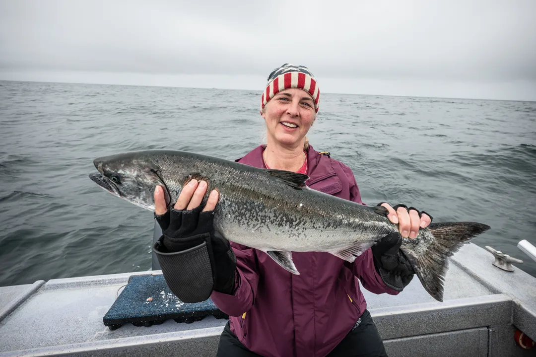 Woman on boat deck holding a silver salmon with open ocean behind her, overcast sky.