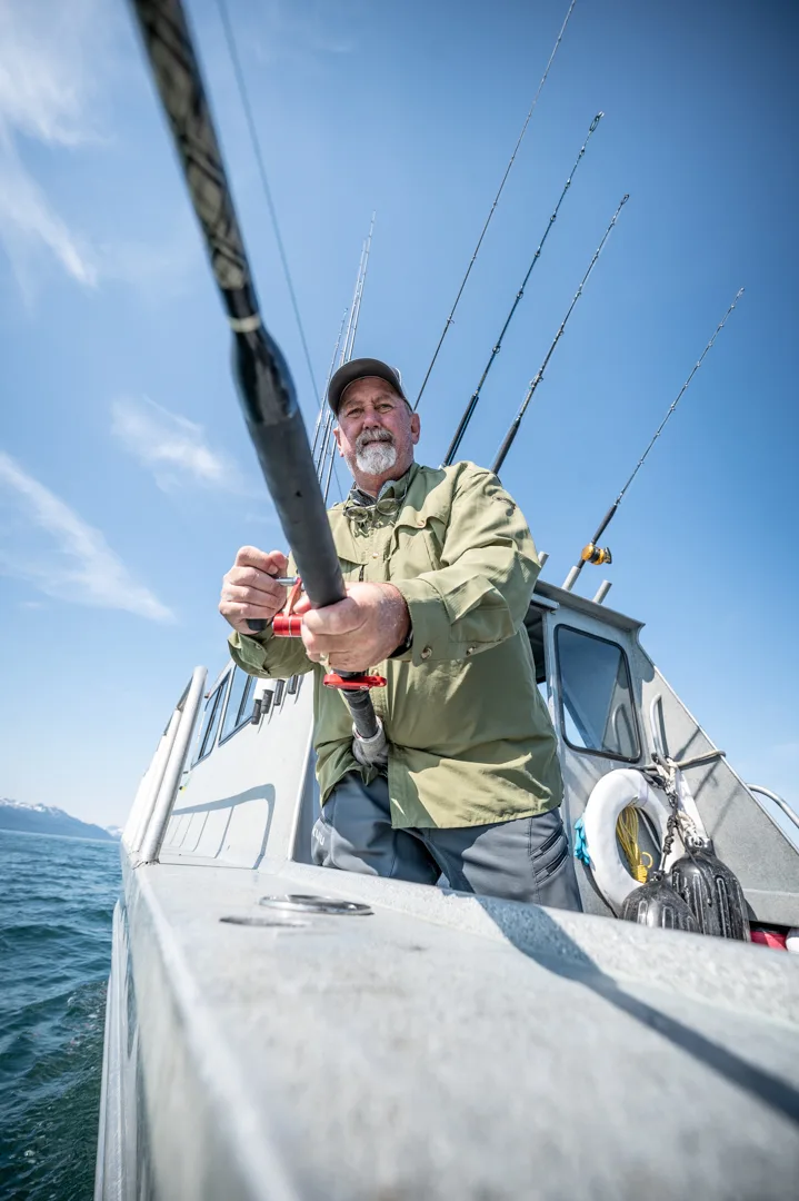 Angler grips a heavy rod on the stern of an aluminum charter boat, multiple rods rigged behind him, blue sky and open water above.