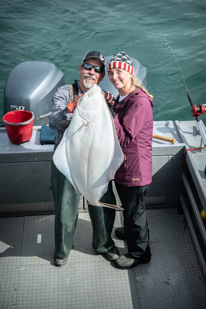 Two anglers holding a large halibut on the deck of an aluminum fishing boat, outboard motors and green water behind them.