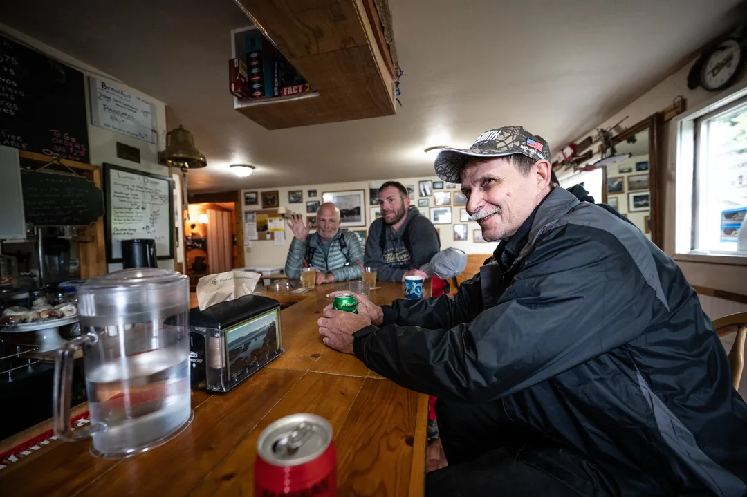 Three anglers relaxing at a wood dining table inside the lodge, chalkboard menus and photos on the walls behind them.