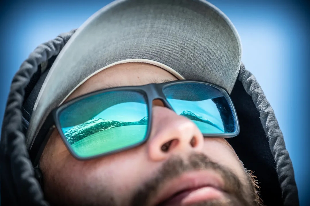 Angler's mirrored sunglasses reflecting snow-capped mountains and glacial water, close-up portrait against blue sky.