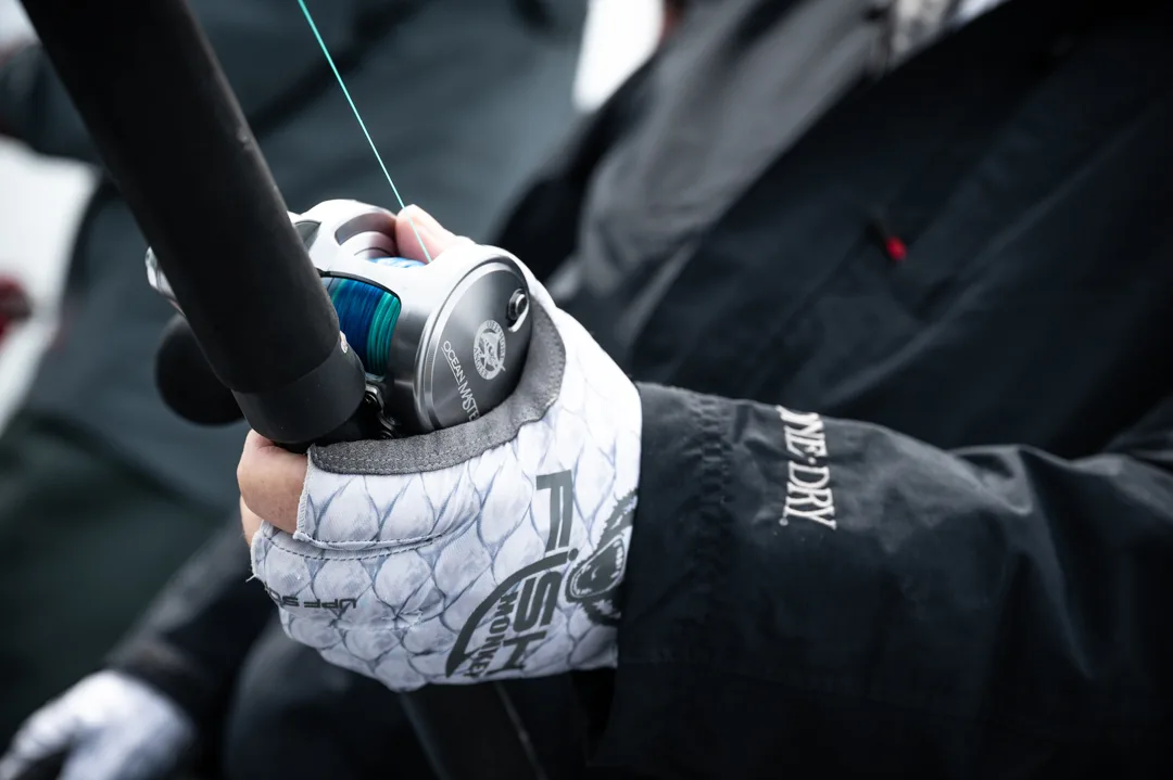 Gloved hand gripping a baitcasting reel and rod on deck, teal braided line loaded on spool.