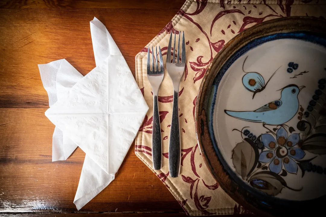 Table setting detail — bird-motif ceramic plate, two forks on floral cloth placemat, folded paper napkin on dark wood.