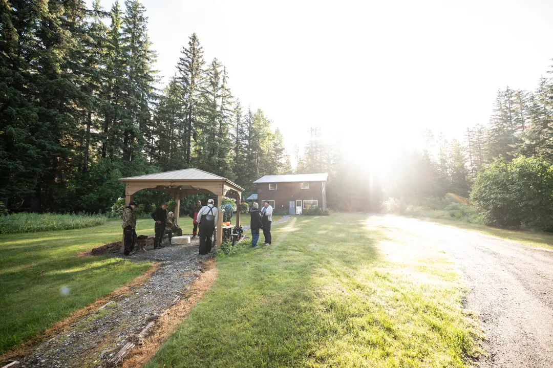 Guests in fishing gear gather under an open pavilion on the lodge grounds at early morning, main building and spruce forest behind.