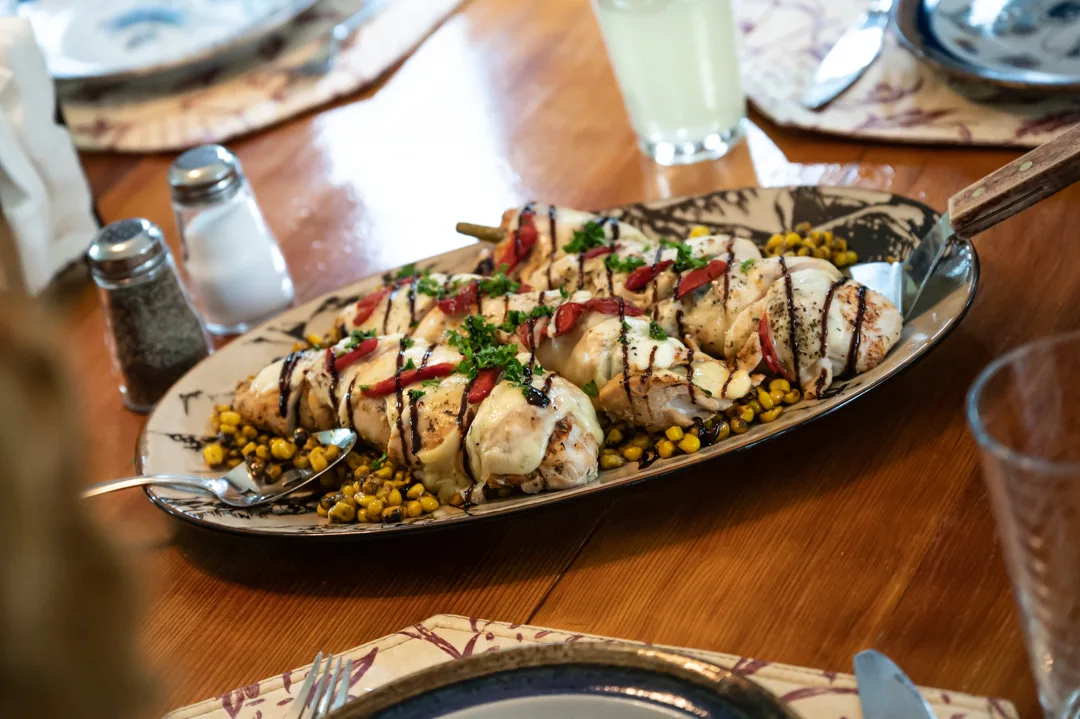 Oval platter of stuffed chicken skewers with roasted corn, red pepper, melted cheese, and balsamic drizzle on a set dining table.